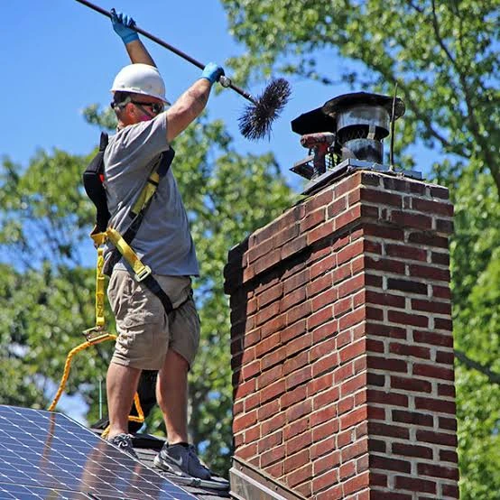 Helmeted person lifting a chimney brush above a brick rooftop chimney